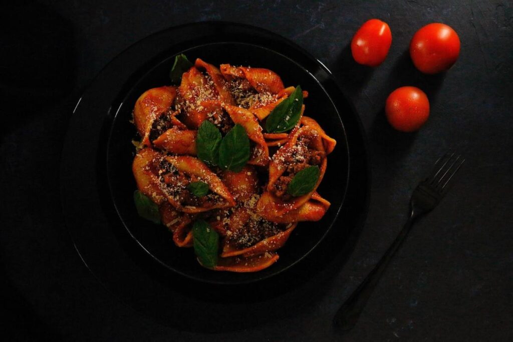 pasta formato conchiglie al ragù con foglioline di basilico e accanto al piatto una forchetta e tre pomodorini